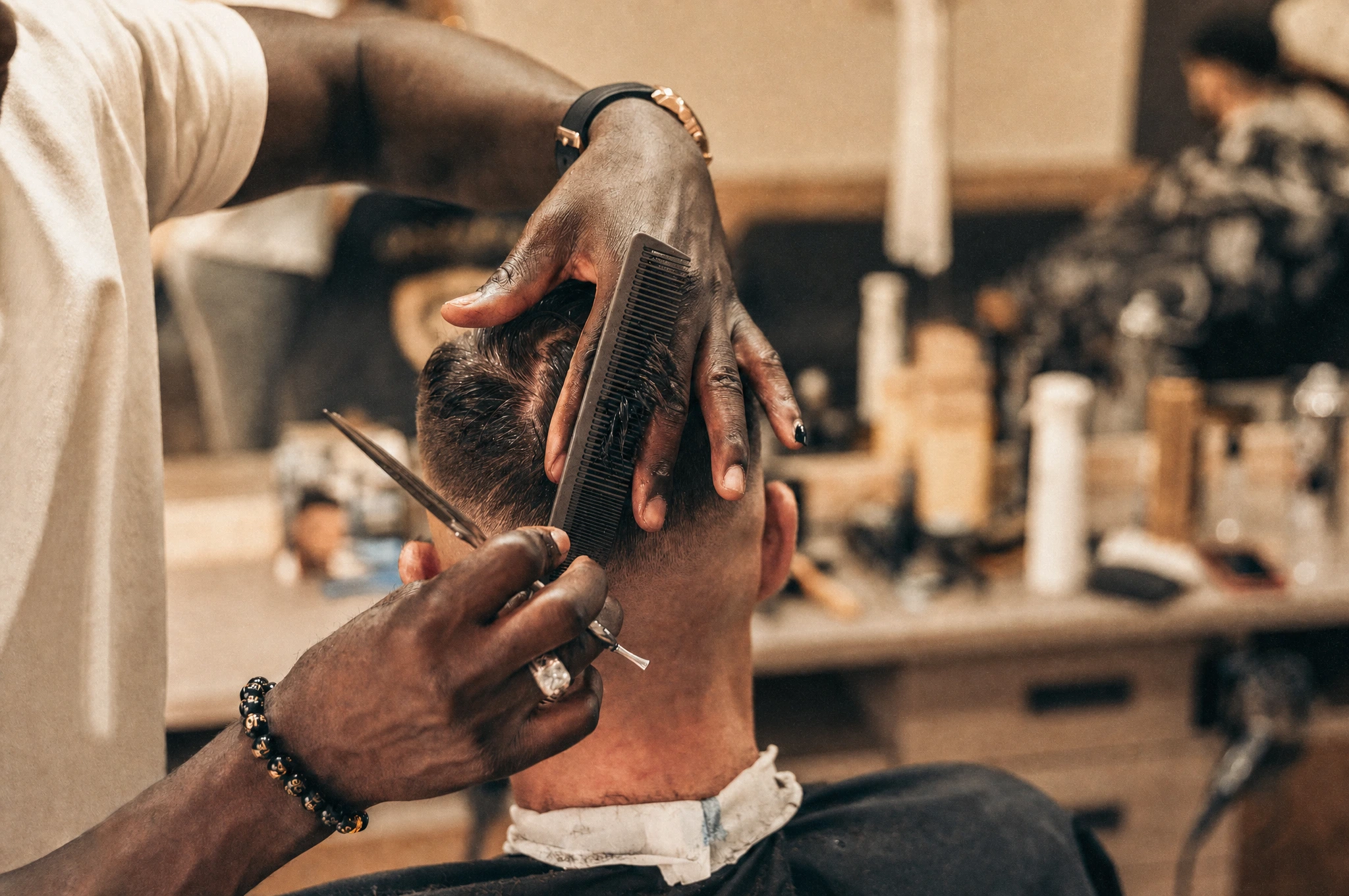 A barber combing and cutting the back of a male client's hair in a warm-lit barbershop.